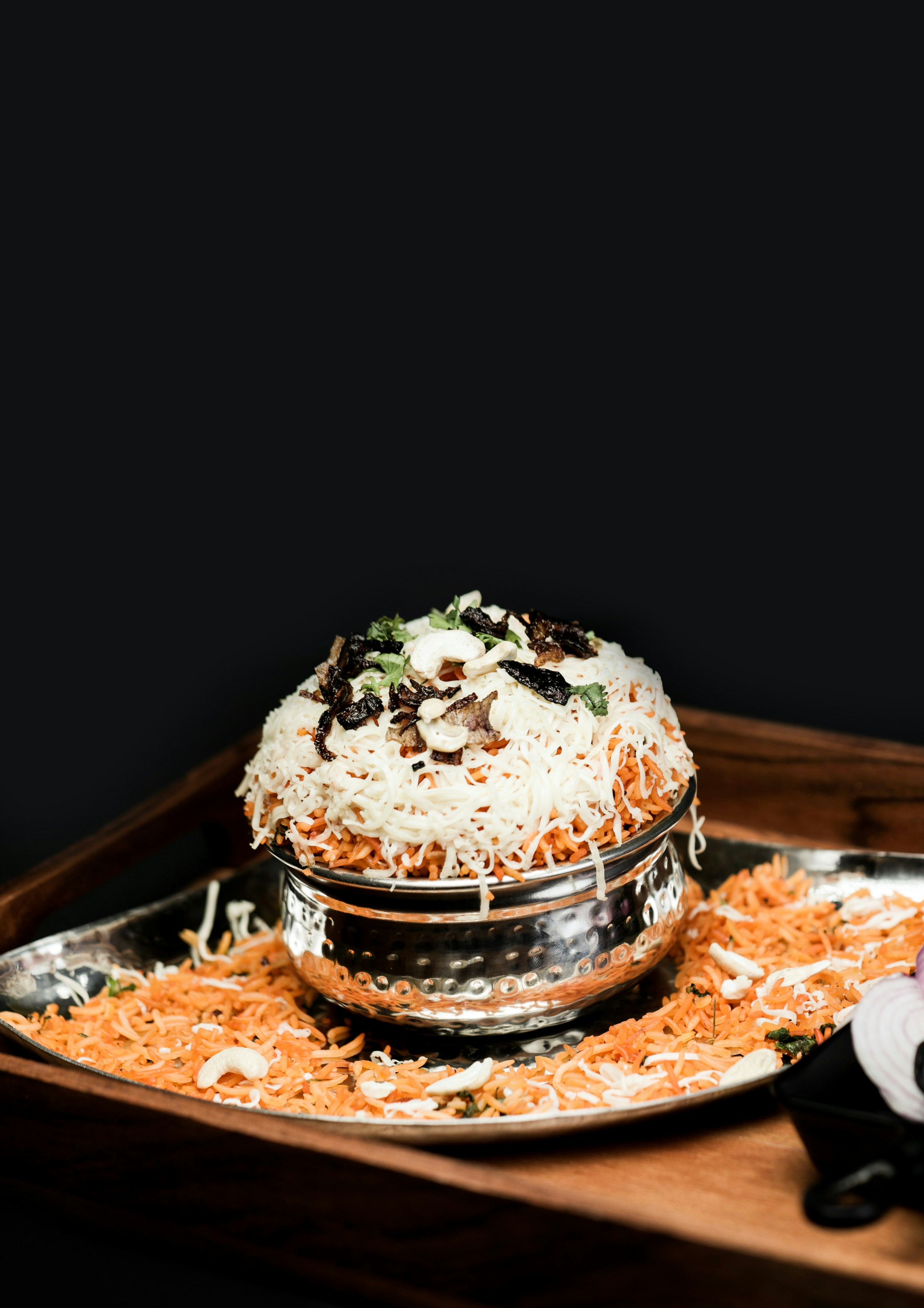 A tall mound of biryani topped with fried onions and nuts, set on a steel pot on a wooden tray
