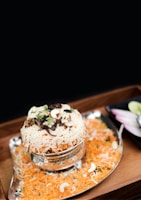 A serving of biryani is presented in a shiny metal bowl, garnished with fresh coriander, crispy fried onions, and cashews. The rice is vibrant with orange hues, featuring layers of spiced biryani rice. The presentation is completed with a silver tray and sliced onions with lime served on the side.