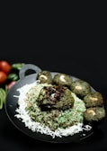 A black platter filled with colorful rice garnished with white shredded elements, topped with a green sauce. There are several green patties with visible cashew nuts encircling the rice, and some fresh vegetables like tomatoes and green beans are partially visible in the background.