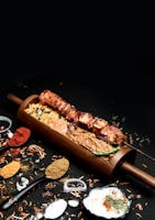 A wooden platter holding skewered grilled meat, surrounded by various colorful spices and herbs. The presentation includes a mix of textures with creamy sauces and vibrant garnishes.
