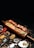 A wooden platter holding skewered grilled meat, surrounded by various colorful spices and herbs. The presentation includes a mix of textures with creamy sauces and vibrant garnishes.