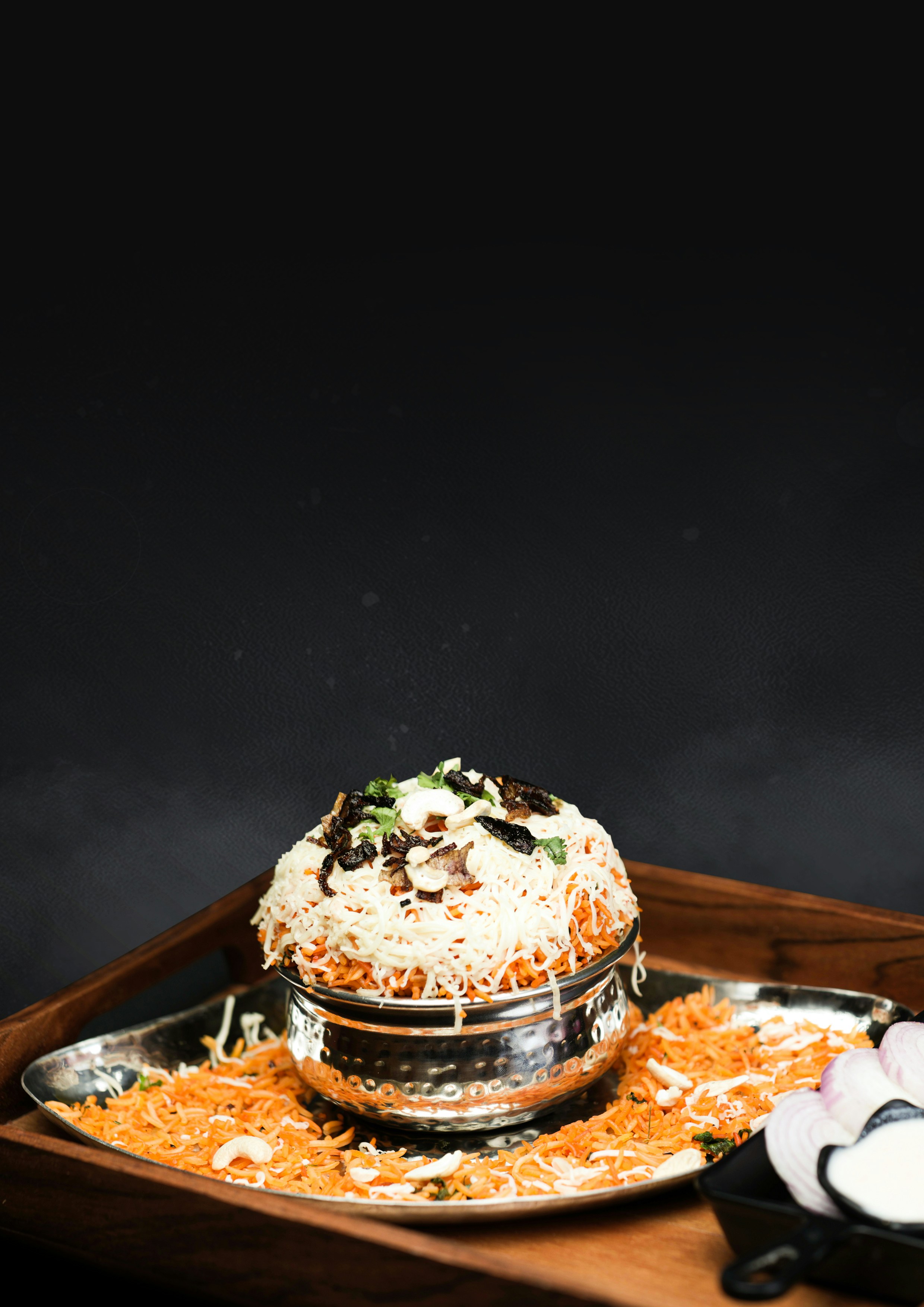 A steel pot of biryani rice garnished with fried onions and nuts, on a steel tray
