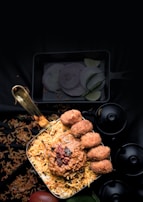 A bowl filled with biryani topped with a spicy sauce and multiple pieces of fried appetizers sits prominently. Surrounding garnishes include sliced onions and green chilies arranged nearby, while scattered grains of rice and a small tomato are visible beneath a dark background.