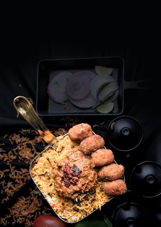 A bowl filled with biryani topped with a spicy sauce and multiple pieces of fried appetizers sits prominently. Surrounding garnishes include sliced onions and green chilies arranged nearby, while scattered grains of rice and a small tomato are visible beneath a dark background.