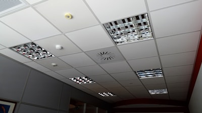 A grid-patterned ceiling with integrated metallic light fixtures, a smoke detector, and ventilation system. The ceiling panels are predominantly white, and the light fixtures consist of mirrored grids. Some ceiling tiles appear slightly darker due to shadowing.