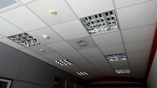 A grid-patterned ceiling with integrated metallic light fixtures, a smoke detector, and ventilation system. The ceiling panels are predominantly white, and the light fixtures consist of mirrored grids. Some ceiling tiles appear slightly darker due to shadowing.