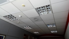 A grid-patterned ceiling with integrated metallic light fixtures, a smoke detector, and ventilation system. The ceiling panels are predominantly white, and the light fixtures consist of mirrored grids. Some ceiling tiles appear slightly darker due to shadowing.