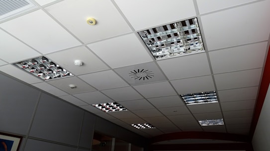A grid-patterned ceiling with integrated metallic light fixtures, a smoke detector, and ventilation system. The ceiling panels are predominantly white, and the light fixtures consist of mirrored grids. Some ceiling tiles appear slightly darker due to shadowing.