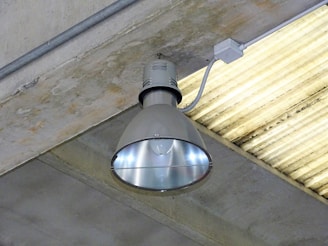 A large industrial-style ceiling light fixture mounted on a concrete ceiling. The lamp has a metallic, reflective surface, and is connected to a power source via a conduit. The surrounding ceiling is made of grey concrete with a linear pattern.