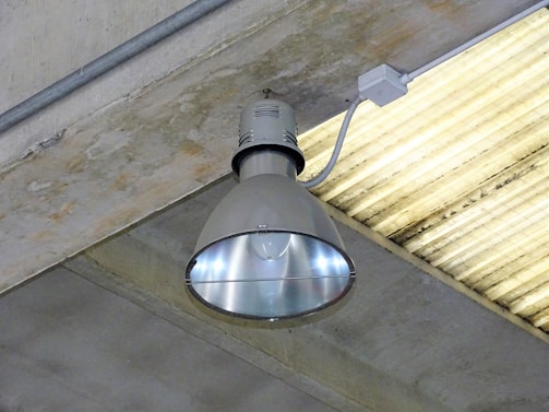 A large industrial-style ceiling light fixture mounted on a concrete ceiling. The lamp has a metallic, reflective surface, and is connected to a power source via a conduit. The surrounding ceiling is made of grey concrete with a linear pattern.