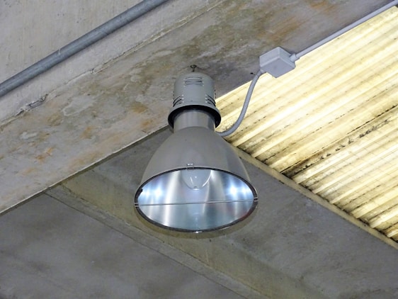 A large industrial-style ceiling light fixture mounted on a concrete ceiling. The lamp has a metallic, reflective surface, and is connected to a power source via a conduit. The surrounding ceiling is made of grey concrete with a linear pattern.