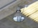 A large industrial-style ceiling light fixture mounted on a concrete ceiling. The lamp has a metallic, reflective surface, and is connected to a power source via a conduit. The surrounding ceiling is made of grey concrete with a linear pattern.