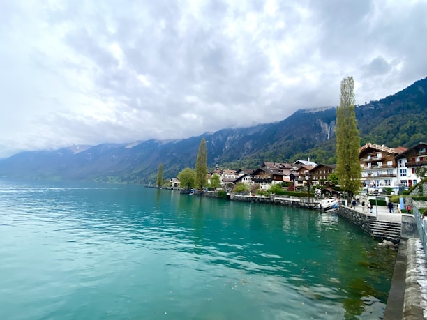 A tranquil lakeside scene with turquoise water stretching out under a cloudy sky. Traditional alpine houses line the water's edge, nestled against a backdrop of lush mountains. Tall, slender trees enhance the picturesque view, while a few people stroll along the stone-paved path by the shore. The landscape exudes a serene and peaceful atmosphere.