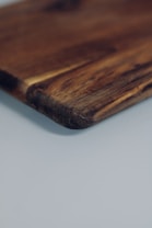 A close-up view of the edge of a wooden cutting board, with a smooth texture and visible grain patterns. The board is positioned against a light, neutral background, enhancing its rich brown tones.