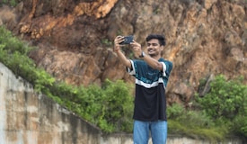 A person is outdoors taking a selfie with a smartphone against a rocky background and lush greenery. The individual is smiling, wearing a casual black and green shirt and blue jeans, and standing on a paved area.