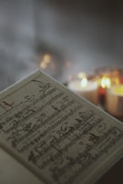 A sheet of music lies open with a vintage or classical look, featuring artistic illustrations amidst the musical notes. In the blurred background, several lit candles provide a warm, soft glow, adding an atmospheric depth to the scene.