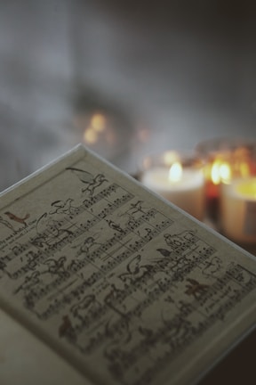 A sheet of music lies open with a vintage or classical look, featuring artistic illustrations amidst the musical notes. In the blurred background, several lit candles provide a warm, soft glow, adding an atmospheric depth to the scene.