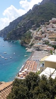 A seaside town with colorful buildings perched on a steep hillside overlooking a vibrant blue sea. The beach is lined with sun loungers and umbrellas, and small boats are scattered across the water close to the shore. Lush greenery and mountainous terrain surround the town.