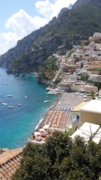 A seaside town with colorful buildings perched on a steep hillside overlooking a vibrant blue sea. The beach is lined with sun loungers and umbrellas, and small boats are scattered across the water close to the shore. Lush greenery and mountainous terrain surround the town.