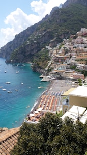 A seaside town with colorful buildings perched on a steep hillside overlooking a vibrant blue sea. The beach is lined with sun loungers and umbrellas, and small boats are scattered across the water close to the shore. Lush greenery and mountainous terrain surround the town.