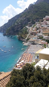 A seaside town with colorful buildings perched on a steep hillside overlooking a vibrant blue sea. The beach is lined with sun loungers and umbrellas, and small boats are scattered across the water close to the shore. Lush greenery and mountainous terrain surround the town.