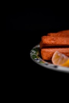A close-up of three slices of golden-brown toasted bread accompanied by a lemon wedge, arranged on a decorative plate with a dark background.