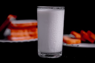 A clear glass filled with a white, opaque liquid, showing condensation on its surface, stands in the foreground. The background features blurred stacks of rectangular slices that are likely orange in color, possibly bread or toast, resting on a plate.