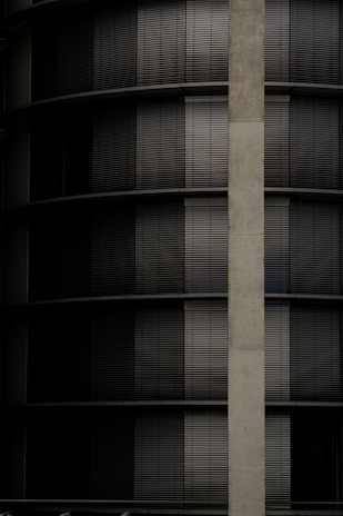A building facade with a series of dark horizontal blinds alternating between closed and partially open positions. A vertical concrete column runs along the right side of the image.