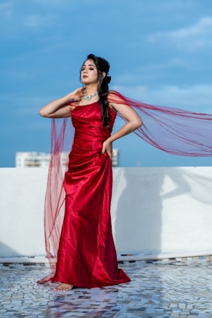A woman stands elegantly on a tiled surface, wearing a flowing red gown with a sheer cape that catches the breeze. Her hair is styled, and she appears to be accessorized with a necklace, embracing a confident posture against a backdrop of a bright blue sky.
