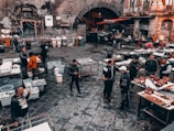 A busy outdoor market scene with people gathered around various tables set with fish, seafood, and other products. The cobblestone ground is wet, indicating a recent rainfall or ongoing cleaning. Surrounding the market are old, worn buildings with graffiti and a large archway leading into a darker area. Vendors and customers, wearing casual clothing, interact and negotiate.