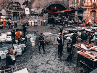 A busy outdoor market scene with people gathered around various tables set with fish, seafood, and other products. The cobblestone ground is wet, indicating a recent rainfall or ongoing cleaning. Surrounding the market are old, worn buildings with graffiti and a large archway leading into a darker area. Vendors and customers, wearing casual clothing, interact and negotiate.