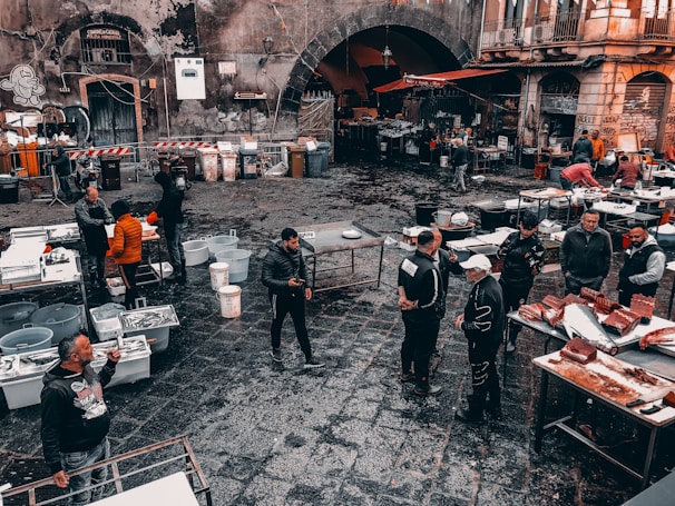 A busy outdoor market scene with people gathered around various tables set with fish, seafood, and other products. The cobblestone ground is wet, indicating a recent rainfall or ongoing cleaning. Surrounding the market are old, worn buildings with graffiti and a large archway leading into a darker area. Vendors and customers, wearing casual clothing, interact and negotiate.