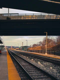 A train platform is situated beside a set of railway tracks extending into the distance. Overhead, an overpass casts shadows on the scene. Sparse vegetation lines the right side of the platform, while the sky is overcast, suggesting an early morning or late afternoon setting.