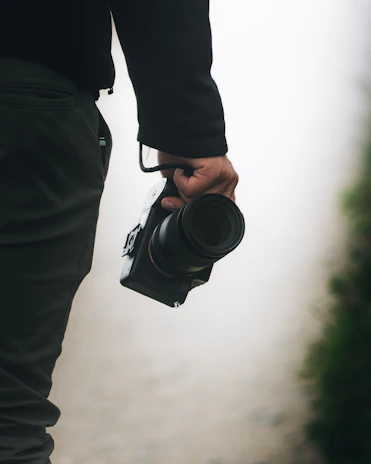 A person holds a professional camera by their side, with their arm extended and the camera strap wrapped around their wrist. The background is softly focused, with hints of greenery and a neutral tone.