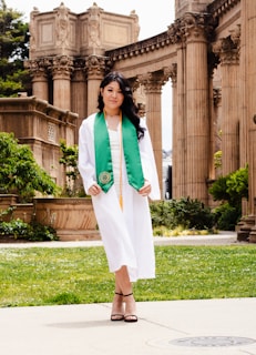 A young woman in a white graduation gown and green sash poses in front of a historic architectural structure with tall columns. The setting appears to be outdoors with greenery and stone pathways.