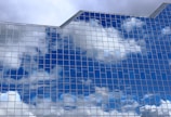 Reflective glass windows of a modern building mirror the blue sky and clouds, creating a pattern of rectangular panes that enhance the architectural aesthetics.