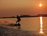 A person is wakeboarding on a calm body of water during sunset. The sun casts a golden reflection on the water, creating a serene and vibrant scene. The silhouette of the wakeboarder adds a dynamic element to the tranquil setting with ripples and splashes visible.