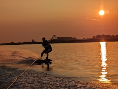 A person is wakeboarding on a calm body of water during sunset. The sun casts a golden reflection on the water, creating a serene and vibrant scene. The silhouette of the wakeboarder adds a dynamic element to the tranquil setting with ripples and splashes visible.