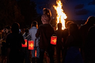 A group of people gather at night, illuminated by a large bonfire. Several individuals are holding red lanterns, some featuring a white cross design. A child is sitting on an adult's shoulders, gazing at the fire.