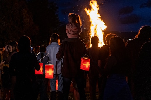 A group of people gather at night, illuminated by a large bonfire. Several individuals are holding red lanterns, some featuring a white cross design. A child is sitting on an adult's shoulders, gazing at the fire.