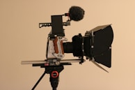 A professional video camera setup is mounted on a tripod. The camera features a matte box, a shotgun microphone with a windscreen, and a side grip with a wood finish. The equipment is composed of multiple intricate components highlighting its advanced functionality and design.