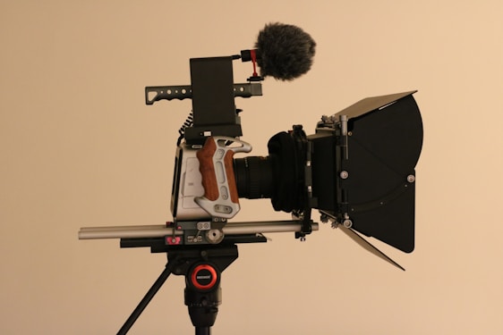 A professional video camera setup is mounted on a tripod. The camera features a matte box, a shotgun microphone with a windscreen, and a side grip with a wood finish. The equipment is composed of multiple intricate components highlighting its advanced functionality and design.