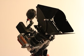 A professional video camera equipped with various accessories is shown. The camera is mounted on a stabilizing rig with a wooden handle, a microphone with a windscreen, and a matte box. The setup appears ready for high-quality video recording.