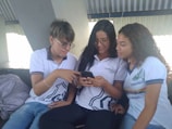 Three young people wearing casual school uniforms are sitting together, engaged with a smartphone. The individuals appear to be sharing something interesting on the phone, and they seem to be in a bright, indoor setting.