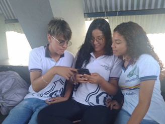 Three young people wearing casual school uniforms are sitting together, engaged with a smartphone. The individuals appear to be sharing something interesting on the phone, and they seem to be in a bright, indoor setting.