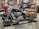 A black motorcycle with leather saddlebags sits on a work platform in a garage, surrounded by tools and automotive equipment. The brick walls and shelves are filled with various mechanical supplies. The motorcycle has shiny chrome details and a tan leather seat and bags with fringe.