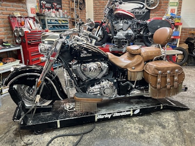 A black motorcycle with leather saddlebags sits on a work platform in a garage, surrounded by tools and automotive equipment. The brick walls and shelves are filled with various mechanical supplies. The motorcycle has shiny chrome details and a tan leather seat and bags with fringe.