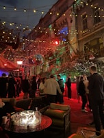 An outdoor social gathering taking place at night, set against a backdrop of a multi-story building with decorative lighting. The area is adorned with string lights and a disco ball, creating a festive atmosphere. A group of people are mingling and chatting, with some seated around a table with a circular fire pit. The lighting gives the scene a warm glow with reds and greens, highlighting the vibrant atmosphere.