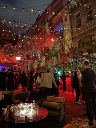 An outdoor social gathering taking place at night, set against a backdrop of a multi-story building with decorative lighting. The area is adorned with string lights and a disco ball, creating a festive atmosphere. A group of people are mingling and chatting, with some seated around a table with a circular fire pit. The lighting gives the scene a warm glow with reds and greens, highlighting the vibrant atmosphere.