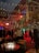 An outdoor social gathering taking place at night, set against a backdrop of a multi-story building with decorative lighting. The area is adorned with string lights and a disco ball, creating a festive atmosphere. A group of people are mingling and chatting, with some seated around a table with a circular fire pit. The lighting gives the scene a warm glow with reds and greens, highlighting the vibrant atmosphere.